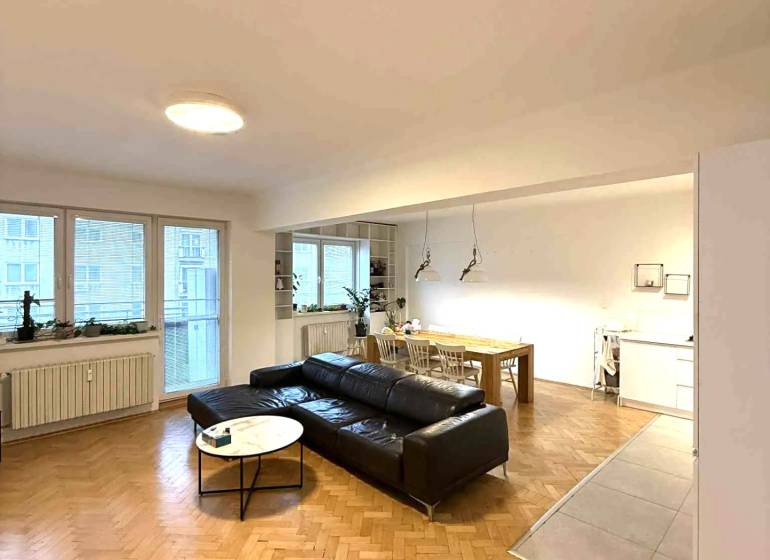 Obývací pokoj ve 4pokojovém bytě s podlahou s dřevěným dekorem a černou pohovkou.