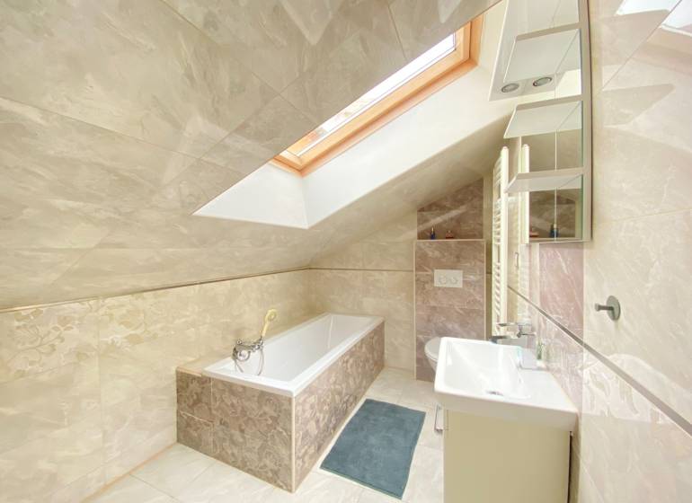 Koupelna v rodinném domě s vanou, oknem ve střeše a světlými obklady.