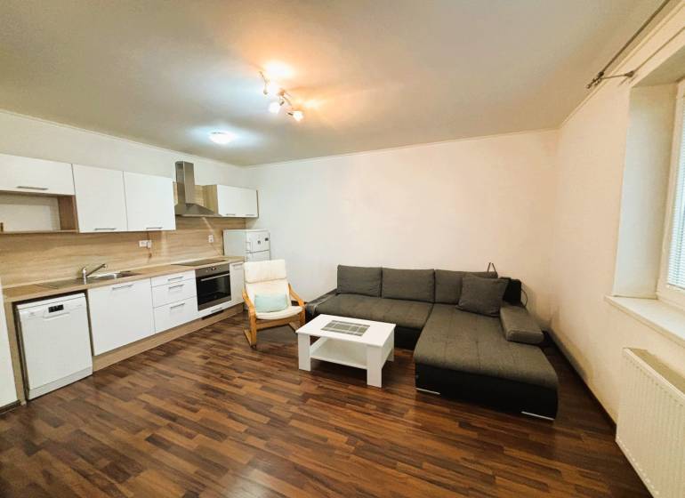 Obývací pokoj v 2-pokojovém bytě s rohovou sedačkou a kuchyňskou linkou.