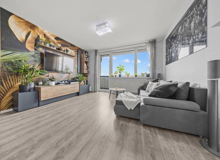 Obývací pokoj s pohodlným gaučem, tapetou s rostlinným motivem a podlahou s dřevěným dekorem.