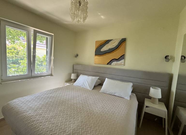 Ložnice v rodinném domě s manželskou postelí, stolky s lampami, velké okno.