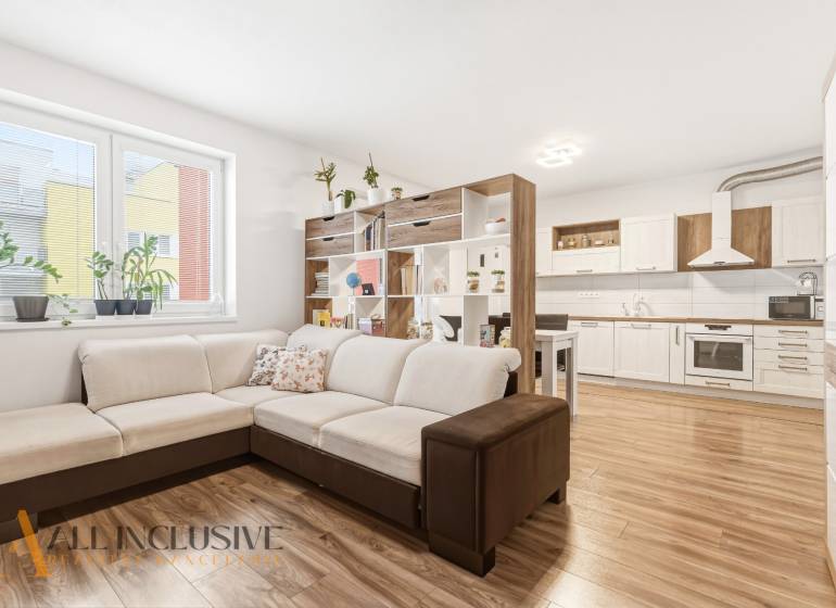 Obývací pokoj v 2-pokojovém bytě s velkou sedačkou a kuchyní, podlaha s dřevěným dekorem.