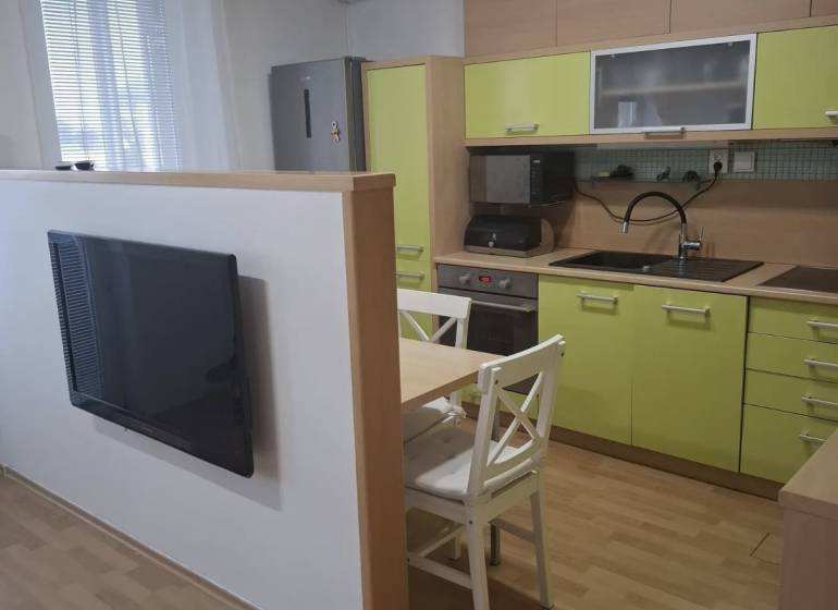 Kuchyně v 2-pokojovém bytě se zelenými skříňkami, televizorem a stolem se dvěma židlemi.