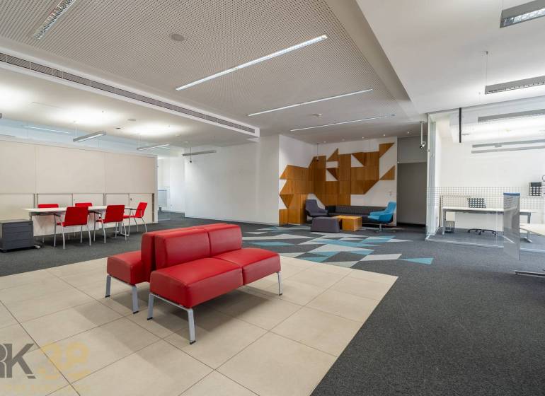 Kanceláře s červenými sedačkami, konferenčním stolem, vzorovaným kobercem a dřevěnými dekoracemi.