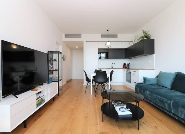 Obývací pokoj s kuchyní v 2pokojovém bytě, podlaha s dřevěným dekorem, TV a sedací souprava.