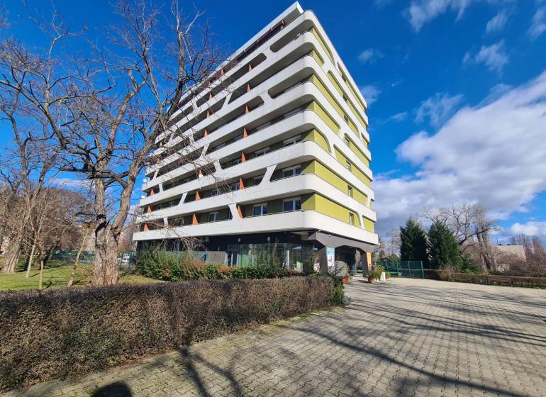 Bytový dům v Komárně se 7 patry, obklopený stromy a dlážděným chodníkem.