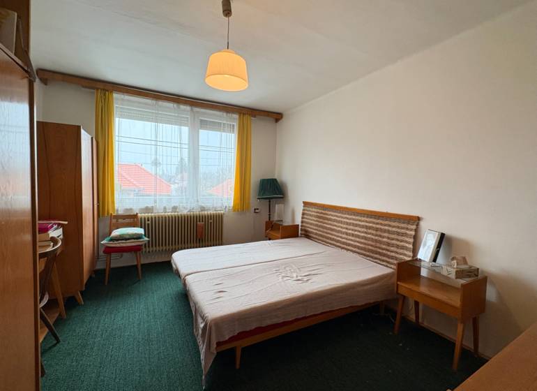 Ložnice s jednoduchým nábytkem v rodinném domě, zelený koberec a žluté záclony.