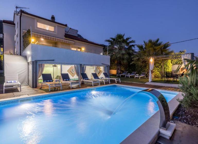 Vila v Kaštela s osvětleným bazénem, lehátky a palmami v zahradě při soumraku.