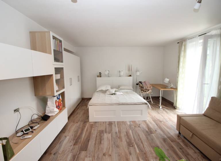 1+kk byt s postelí, sedačkou a skříní, podlaha s dřevěným dekorem.