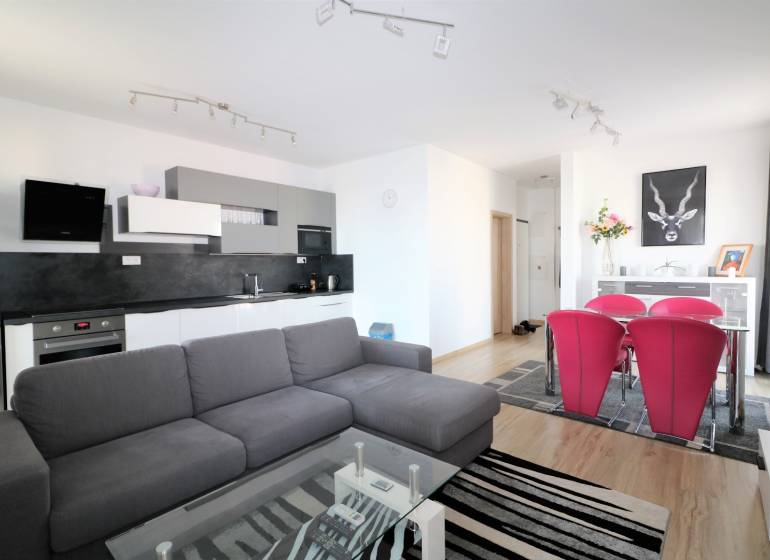 Obývací pokoj s kuchyňským koutem, šedou sedačkou, skleněným stolem a červenými židlemi.