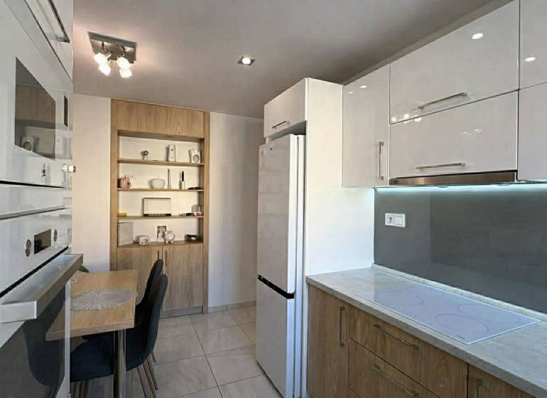 Kuchyně v 2-pokojovém bytě s bílými skříňkami, stolem a dřevěným dekorem polic.