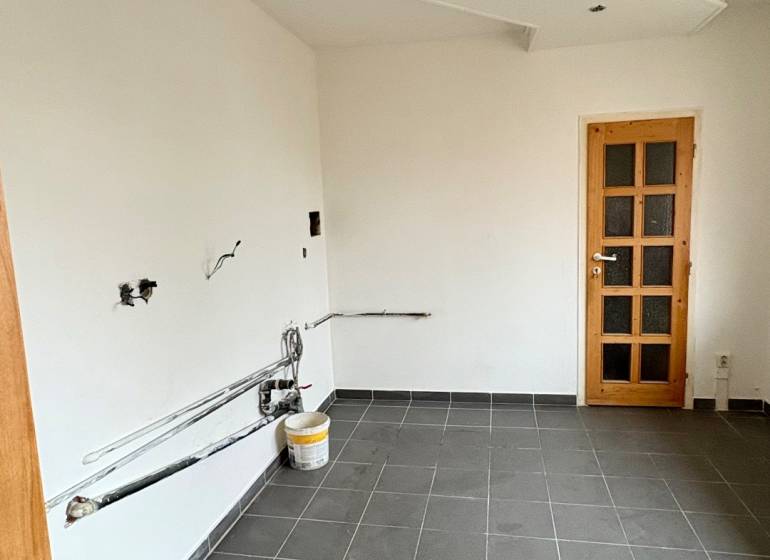 Prázdná místnost v 2-pokojovém bytě, podlaha z tmavé dlažby, bílé stěny, dřevěné dveře.