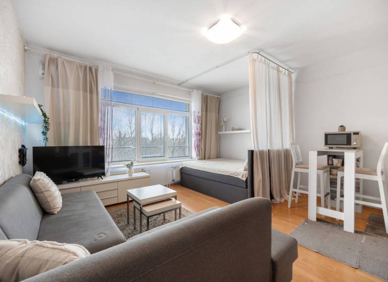 Obývací část 1-pokojového bytu s pohovkou, TV, jídelním stolem a postelí.