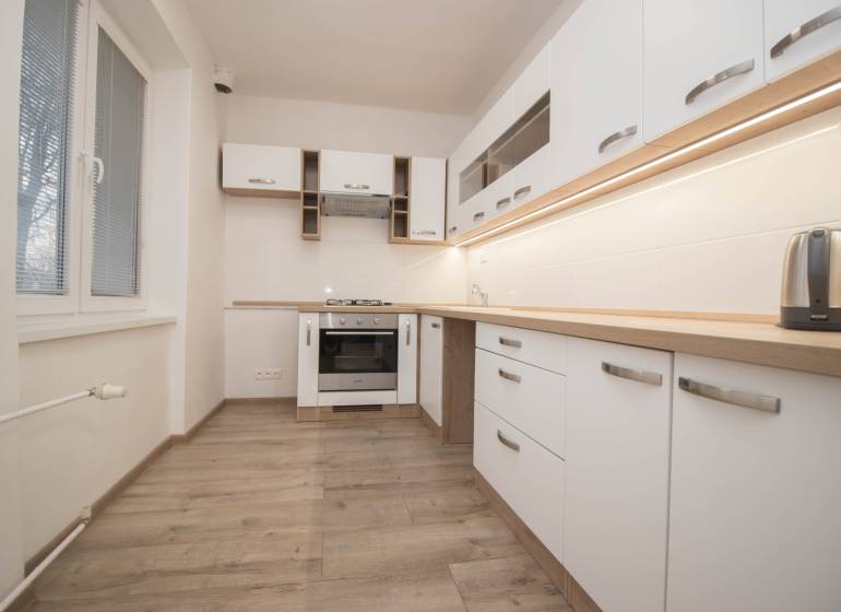 Kuchyně se spotřebiči, bílými skříňkami a podlahou s dřevěným dekorem v 2-pokojovém bytě.