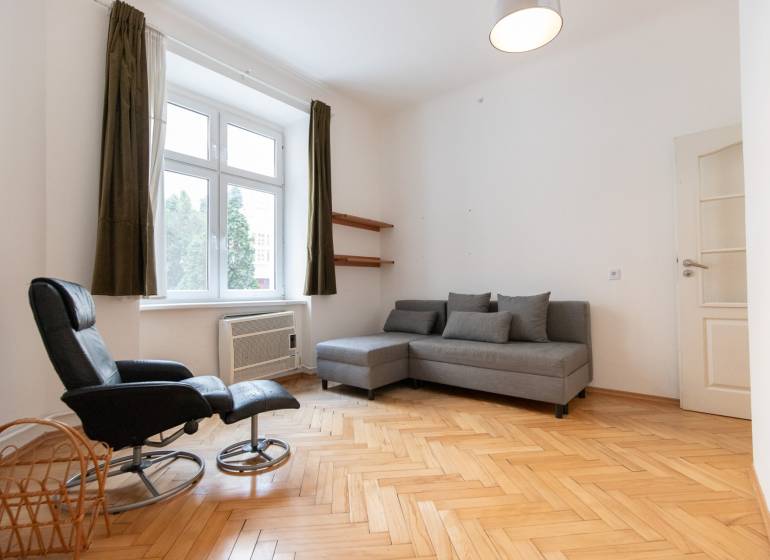 Světlý obývací pokoj s podlahou s dřevěným dekorem ve 4-pokojovém bytě, šedá sedačka, křeslo.