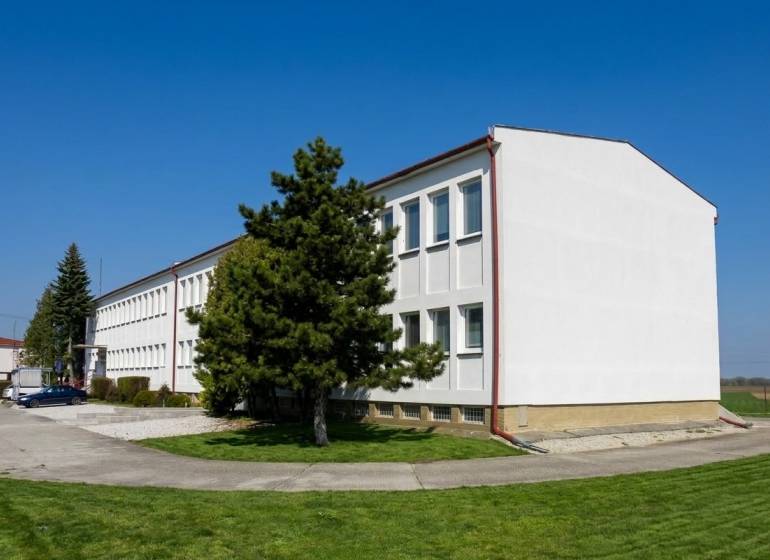 Budova v Šoporni obklopená zelení a stromy, s jasnou modrou oblohou.