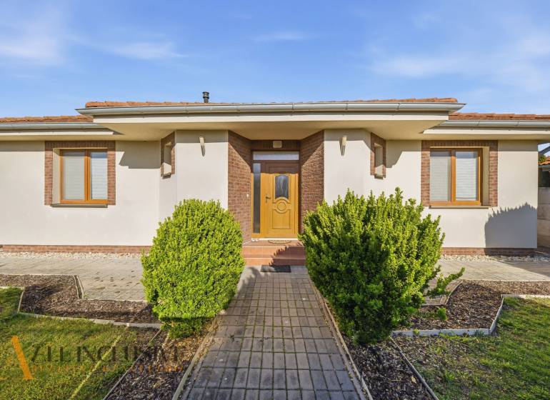 Rodinný dům v Kútnikách s keřovou výsadbou, bílou fasádou a hnědými doplňky.