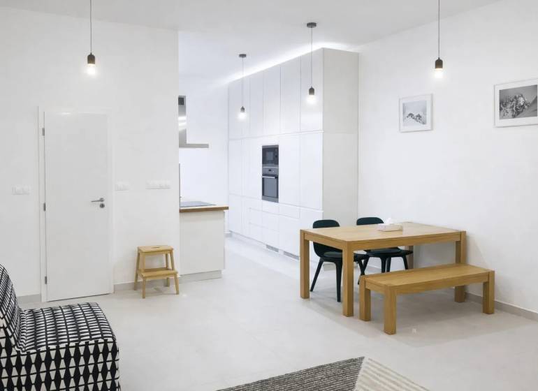 Interiér 2-pokojového bytu s bílými stěnami, dřevěným stolem a černou sedačkou.