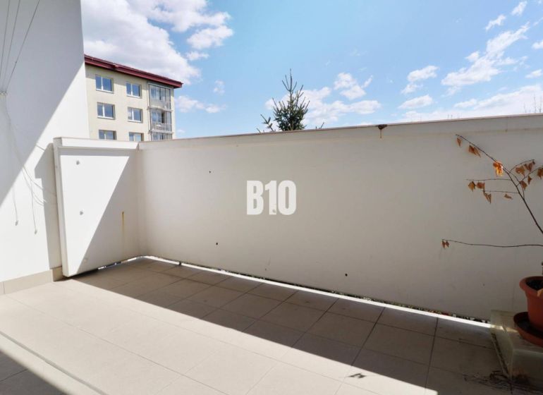 Balkon v 3-pokojovém bytě. Rostlina v květináči, dlažba, částečný výhled na sousední budovu.