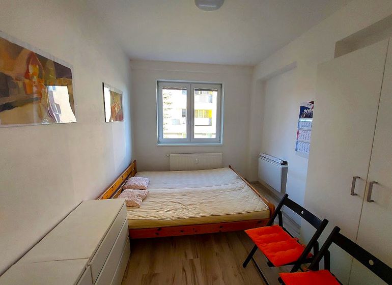 Garsonka s postelí, bílou skříní a podlahou s dřevěným dekorem, dvě sklápěcí židle.