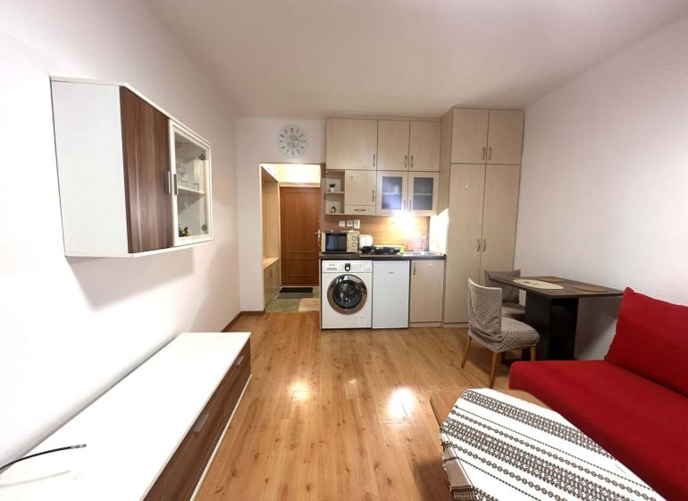 Rekreační apartmán s vestavěnou kuchyní, červenou sedačkou a podlahou s dřevěným dekorem.