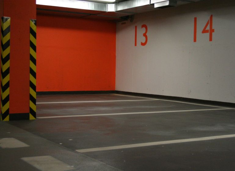 Parkovací místa a oranžová zeď v garáži se žlutými a černými sloupy.