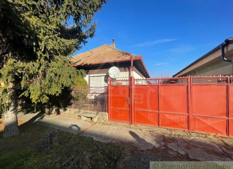 Červená brána a plot s rodinným domem v Tvrdošovcích, obklopený zelení.