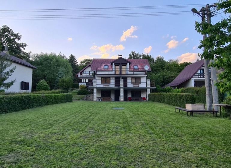 Chata v Kalúži obklopená zelení, udržovaný trávník, okolí s dřevinami, modrá obloha.