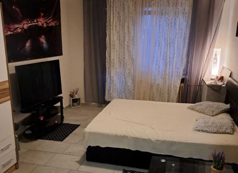 Garsonka s šedo-bílými stěnami, postelí, televizí a dlažbou na podlaze.