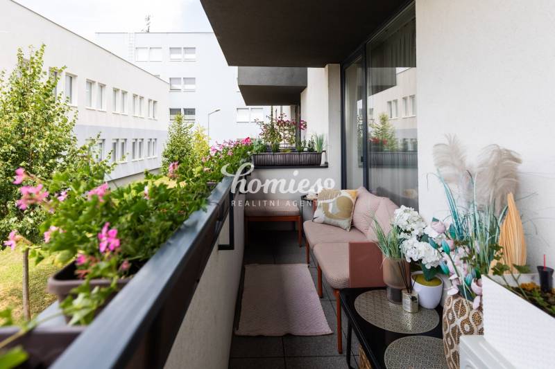 Balkon 2pokojového bytu s květinami, pohovkou a výhledem na okolní budovy.