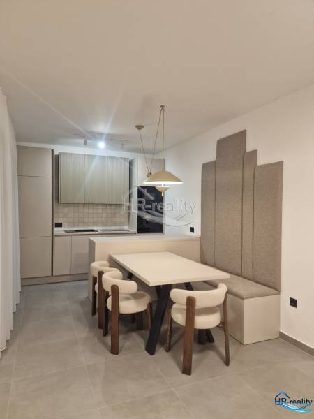 Jídelna v rekreačním apartmánu s kuchyní, světlými židlemi a šedými panely na stěně.