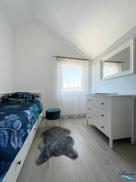 Ložnice v rekreačním apartmánu s postelí, bílou komodou a křeslem na dřevěné podlaze.