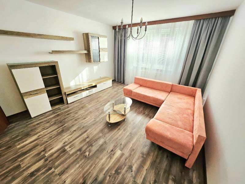 Obývací pokoj třípokojového bytu s rohovou sedačkou, závěsy a dřevěnou podlahou.