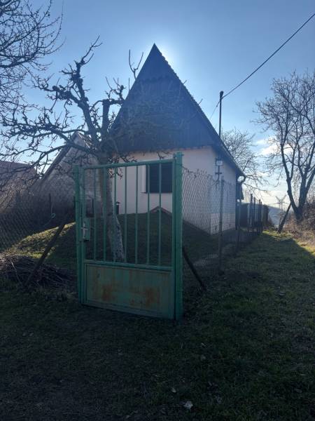 Malá chata obklopená stromy, oplocená zahrada, ideální pro rekreační pozemky.