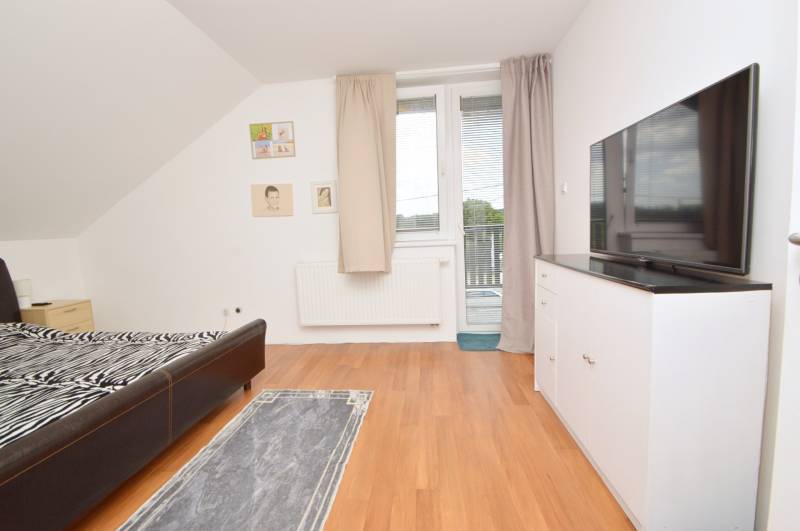 Ložnice v rodinném domě s postelí, televizí, skříňkou a přístupem na balkon.