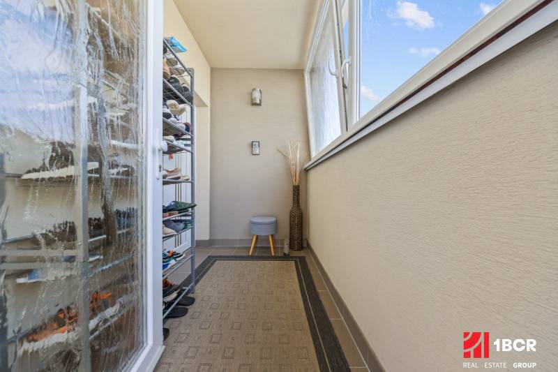 Balkon ve 4-pokojovém bytě s botníkem, koberec, zasklení, otevřené okno.