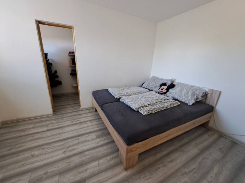 Ložnice rodinného domu s postelí, vínovou podlahou a vchodem do komory.