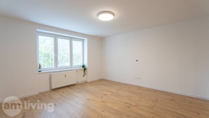 Prostorný pokoj s velkým oknem a dřevěnou podlahou v 1-pokojovém bytě.
