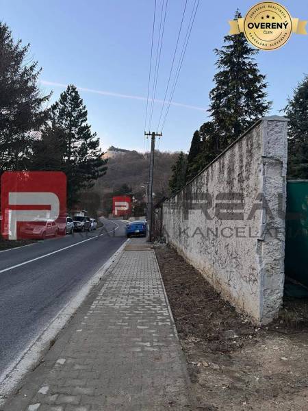Cesta s chodníkem vedoucím přes Pezinok u rodinného domu, v pozadí kopcovitá krajina.