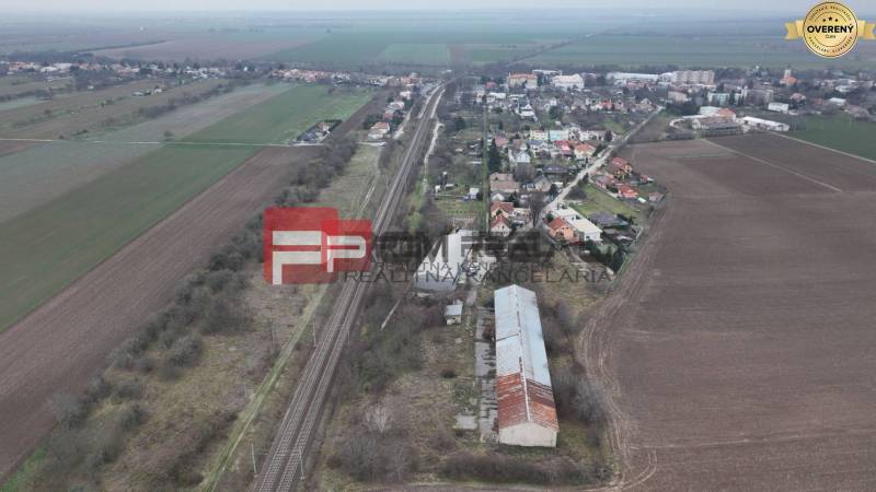 Železniční trať obklopená poli a domy, na ulici Partizánska, jsou viditelné sklady a haly.