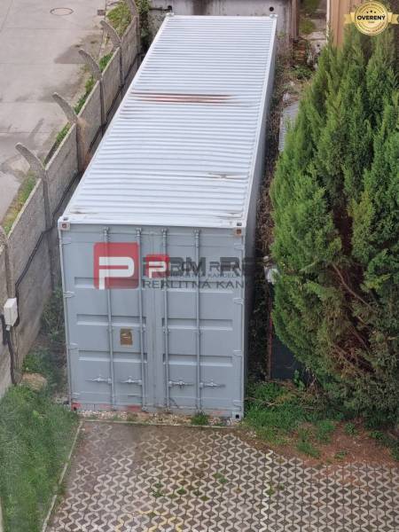 Na Púchovské ulici v Bratislavě III je lodní kontejner vedle zeleně a plotu.