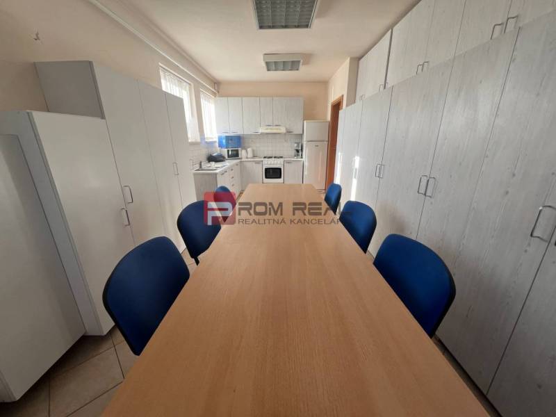 Kuchyňka ve výrobních prostorách s dřevěným stolem, modrými židlemi a bílými skříňkami.