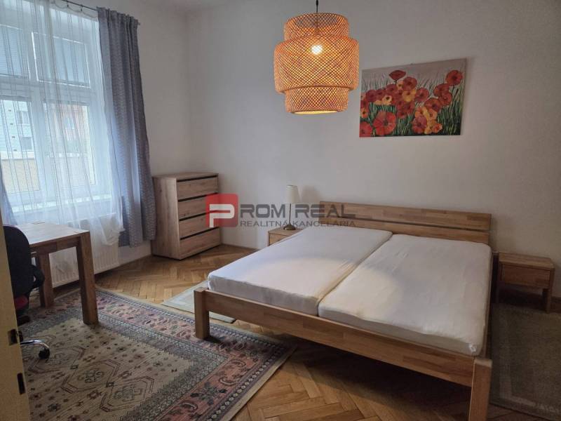 Ložnice s dřevěnou postelí, pracovním stolem a světlým dekorem v 2-pokojovém bytě.