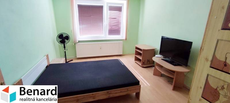 Ložnice v dvoupokojovém bytě s postelí, televizorem a ventilátorem v rohu místnosti.