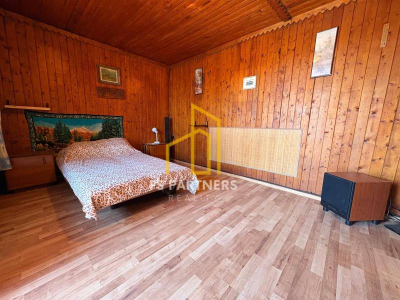 Dřevěný interiér s postelí, dekorativní tkanina na stěně, obrazy a jednoduchý nábytek.