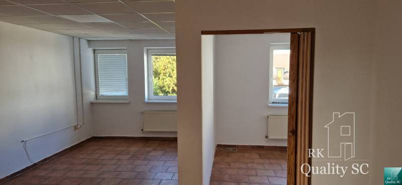 Interiér budovy s bílými stěnami, hnědou dlažbou a třemi okny s výhledem.