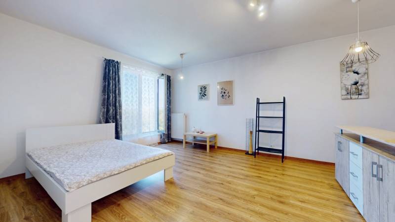 Prostorný pokoj s postelí, dřevěnou podlahou, nástěnnou dekorací a velkým oknem v 1-pokojovém bytě.