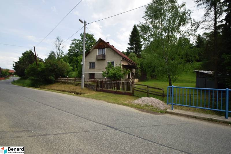 Rodinný dům ve Velké Lesné u cesty, obklopený zelení a plotem.