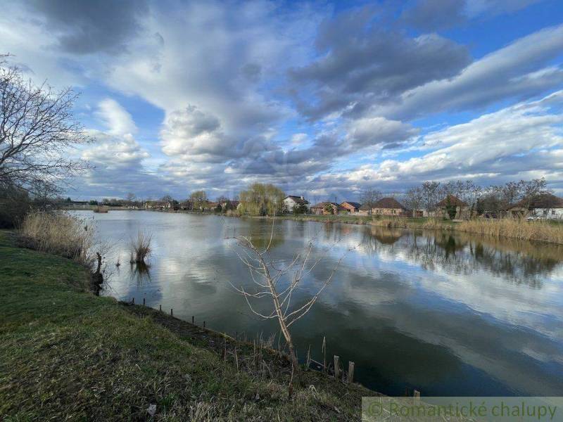 Idylická krajina s jezerem, oblaky a rodinnými domy v pozadí u břehu.