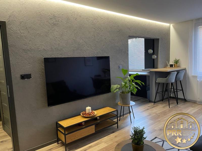 Obývací pokoj v 3-pokojovém bytě s moderní TV, rostlinami a barovým pultem s vysokými stoličkami.
